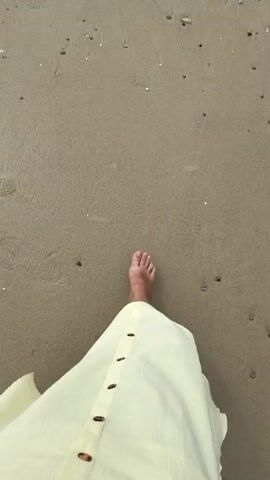This video shows a first-person perspective walking on a sandy beach. The camera pans upwards, revealing the ocean and a rocky shoreline. The sand is light brown with small pebbles and shells scattered across it. In the foreground, a white garment, possibly a dress or skirt, is visible. Waves are gently rolling in and breaking against the rocks. The sky is partly cloudy with a mix of blue and white. There are a few indistinct figures visible on the distant rocks. The overall scene is peaceful and serene.
