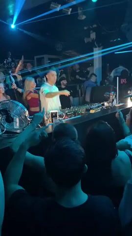 The video shows a DJ performing in a nightclub. The DJ is standing behind a console with turntables and a mixer, wearing a white t-shirt and dark pants. Blue and green laser lights are cutting through the dark room, illuminating the crowd. People are dancing and holding drinks. A large fan is positioned on the left side of the DJ booth. The atmosphere appears energetic and festive.