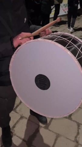 Here's a description of the video:

The video shows a group of people outdoors at night. The focus is on a person holding a large drum and drumsticks. The drum has a white head with a black circle in the center and a dark-colored frame with a lattice design. The person is about to strike the drum. In the background, a group of people is holding up a banner. They appear to be standing on a brick or stone-paved area. The lighting is dim.