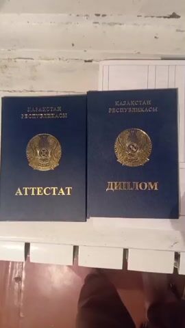 A person's hand is shown in the video, as it is opening a blue booklet with a golden emblem on the cover. The booklet has the words "KAZAKHSTAN REPUBLICASY" and "ATTESTAT" written on it in gold lettering. The booklet appears to be a document or certificate. The background is out of focus but appears to be a wooden surface.