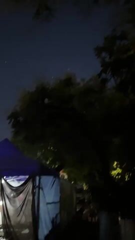 A nighttime video shows a dark outdoor scene with trees in the foreground and fireworks exploding in the sky. A purple tent is visible on the left side of the frame, and other tents or structures are behind it. The fireworks are bright and colorful, with sparks and smoke filling the sky. The trees are silhouetted against the dark sky, and their leaves are illuminated by the fireworks. The video is shot from a low angle, looking up at the sky.