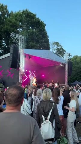 A live music performance is taking place outdoors on a stage with a large crowd gathered in front of it. The stage is set up with a white canopy, lighting rigs, and speakers. Bright pink and white lights illuminate the stage, where a female singer in a white outfit is performing, accompanied by musicians. The crowd, a mix of adults and young people, is facing the stage, some appearing to be filming the performance. The scene is set in the evening, with a clear blue sky visible above the trees in the background. The overall atmosphere seems to be one of enjoyment and excitement. The location is identified as Panevėžys, Lithuania.