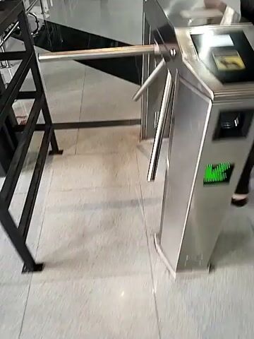The video shows a first-person perspective of someone walking towards and through a turnstile. The turnstile is made of brushed metal and has a horizontal bar that rotates to allow passage. The floor is tiled with a pattern of light and dark squares. In the background, there is a black and white checkered floor. The person is wearing black sandals. The video is shot in landscape orientation.