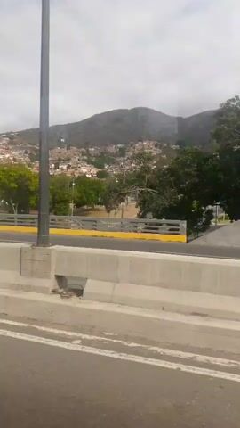 The video shows a view from a moving vehicle, likely on a highway, in a somewhat urban or suburban area with a backdrop of hills or mountains. The sky is overcast. Alongside the highway, there is a grassy area with trees and a road. A couple of people are seen riding a small motorcycle or scooter on the side road, and a small trailer towed by a motorized vehicle passes by. The video passes a highway sign. The focus is primarily on the roadside scenery and the vehicles on the adjacent road.