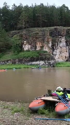 The video shows a wide, calm river with muddy brown water. The riverbank in the foreground is covered with grass and gravel, and there is an orange inflatable raft with paddles, bags, and other gear loaded onto it, suggesting it is prepared for use. In the background, a steep, rocky cliff face covered with trees rises from the opposite bank. Several people are visible on the river in various small boats, including kayaks and what appear to be rafts, all moving downstream. Some people are standing on the riverbank, observing the activity. The sky is overcast, and the overall impression is of a recreational outing on a river surrounded by natural scenery.