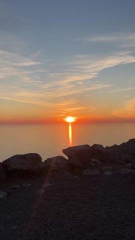 The video depicts a sunset over a body of water, likely the ocean. The sun is low on the horizon, casting a bright orange and yellow glow across the sky and water. Thin, wispy clouds are visible in the upper part of the frame, colored with the orange and pink hues of the setting sun. In the foreground, there are rocks and gravel, silhouetted against the bright light. The composition is static, with no visible camera movement or zooming. There are no people visible in the shot.