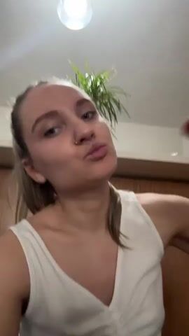 A woman is eating a slice of red apple in a room with a plant and wooden furniture. The woman has blonde hair pulled back into a ponytail, a nose ring, and red nail polish. She wears a white tank top and looks directly at the camera between bites, chewing with her mouth closed. The lighting is bright, and the camera angle is slightly upward, as if the viewer is shorter than the subject. The background is softly focused.