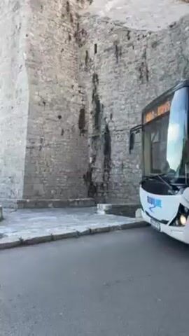 The video shows a white bus with black accents, emblazoned with "BLUELINE" on its front, slowly moving along a paved road. To the left, a large, ancient-looking stone wall or building, possibly a fortress or castle, dominates the background. The wall is constructed from light-colored, rough-hewn stone blocks.

As the bus pulls forward, the scene opens up to reveal a picturesque coastal view. A vast expanse of blue water, likely the sea, stretches to the horizon, meeting distant hills or mountains under a clear blue sky with sparse clouds.

At the 0:01 mark, a woman in a light-colored coat is seen near a black stroller on the sidewalk, positioned in front of the stone wall. Another person is partially visible next to her. Further along the sidewalk, at the 0:02 mark, two women are sitting on stone steps or a low stone wall. One woman is dressed in a gray top and light-colored pants, looking at her phone. The other, wearing a dark jacket and white pants, carries a backpack.

The setting appears to be a sunny day. A tall streetlamp post and a signpost with multiple directional signs (featuring red, green, and blue panels) are visible. A white crosswalk is painted on the asphalt. In the background, there are green bushes, trees, and some palm trees. A white trash or recycling bin is also visible with the text "ČISTOĆA" and "21. - 06." on its side, suggesting a cleaning service or schedule. The bus continues to move past the seated women and the street signs before the video ends.