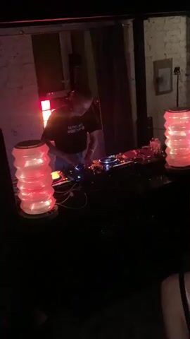 A DJ is mixing music in a dimly lit space. The room has brick walls. The DJ stands behind a table covered in equipment, flanked by two stacked lamps that emit a warm light. A grid of lights behind the DJ flashes in patterns.  The DJ is wearing a black t-shirt with white lettering.  A small portion of a person's head is visible on the right side of the frame.