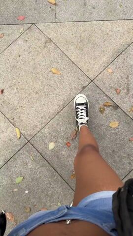 The video shows a first-person view of someone walking on a paved walkway. The person is wearing blue denim shorts and black and white Converse sneakers. The walkway is made of gray stones arranged in a herringbone pattern, with some sections being larger, smooth gray slabs. Scattered on the walkway are a few fallen leaves in various shades of yellow, orange, and red. The camera angle is looking down towards the feet as the person walks.