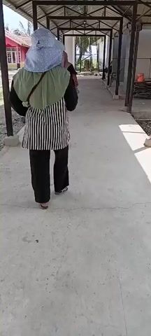 TWO PEOPLE WALK DOWN A COVERED WALKWAY. THE PERSON IN THE FOREGROUND IS WEARING A LIGHT BLUE HAT WITH WHITE DOTS, A GREEN HIJAB, AND A BLACK AND WHITE VERTICALLY STRIPED SHIRT OVER BLACK PANTS. THEY ARE WEARING SANDALS. THE PERSON IN THE BACKGROUND IS WEARING A DARK HIJAB AND A DARK DRESS. THEY APPEAR TO BE HOLDING A METAL ROD. THE WALKWAY HAS A CONCRETE FLOOR AND IS COVERED BY A METAL ROOF SUPPORTED BY METAL POSTS. TO THE LEFT OF THE WALKWAY IS A PINK BUILDING WITH A RED ROOF AND SOME TREES. TO THE RIGHT OF THE WALKWAY ARE SOME CONSTRUCTION MATERIALS, INCLUDING WHAT APPEAR TO BE LARGE, FLAT BLOCKS. THE GROUND IS DUSTY AND UNEVEN IN PLACES. THE SKY IS BRIGHT AND APPEARS TO BE CLOUDY.