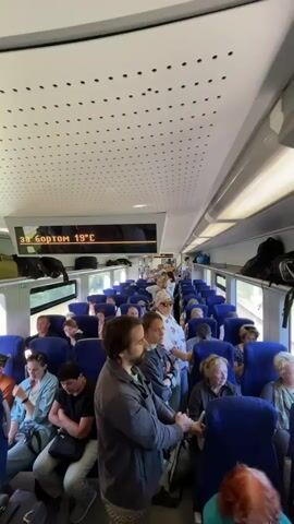 The video shows the interior of a train carriage filled with passengers. The camera is positioned in the aisle, looking forward down the length of the carriage. Blue upholstered seats are arranged in rows on either side of the aisle, most of which are occupied by people. Passengers are of various ages and genders, some are looking around, others are looking down or at their phones. Luggage is visible on overhead racks. A digital display above the aisle shows "ЗА БОРТОМ 19°C", indicating the outside temperature is 19 degrees Celsius. The lighting is bright, suggesting daytime. Some passengers are standing in the aisle. The overall impression is of a moderately crowded train journey.
