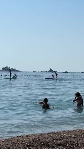 This is a vertical, medium shot video of a beach scene with people swimming and paddleboarding. In the foreground, two people are swimming in the ocean. One person, wearing dark swimwear, is facing away from the camera and splashing water with their right hand. Next to them, a child with light-colored swimwear is looking towards the shore. To the right, partially obscured by the water, a person in a bikini is visible. In the mid-ground, several people are paddleboarding. On the left, a person stands on a paddleboard and is facing the shore. To their right, another person sits on a paddleboard. In the center of the mid-ground, two individuals are on a paddleboard, one standing and paddling, the other sitting. Further in the background, more paddleboarders are visible. In the far distance, islands with greenery can be seen under a clear blue sky. The water is calm with gentle waves lapping at the pebble beach in the foreground.