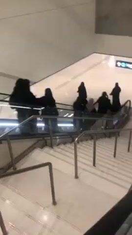 The video shows an escalator in a modern, brightly lit building. Several people, most dressed in black, are riding the escalator up. There are additional stairs with metal handrails alongside the escalator. A directional sign is visible overhead. The building appears clean and minimalist in design.