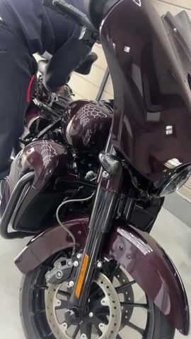 A person, wearing dark clothing with a red stripe on the leg, is leaning over a dark purple motorcycle. The motorcycle has a glossy finish with a hexagonal pattern visible on the fuel tank and fairing. The front wheel with disc brakes and suspension is also visible. The scene appears to be indoors, possibly a workshop or garage, with wooden paneling on the wall in the background. There are no strong indicators of time of day or weather. The overall mood is neutral, focusing on the activity of inspecting or working on the motorcycle. No visible text is present on the motorcycle or in the environment.