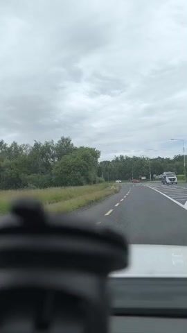 The video captures a forward-facing view from inside a vehicle, likely a car or RV, as it travels along a road in a rural setting. The scene is dominated by a gray, overcast sky, suggesting a cloudy or possibly rainy day. Lush green trees and vegetation line both sides of the road, with a strip of grass and wildflowers separating the asphalt from the dense foliage on the left.

The road itself is paved, with clear yellow and white lane markings. The vehicle is moving on a section of road that curves to the right. In the distance, a white car is visible, as are streetlights and signs. A white RV is parked on the right side of the road near an intersection or junction. The perspective is slightly obstructed by the interior of the vehicle in the foreground, specifically what appears to be a black dashboard accessory or camera mount.

The overall mood is neutral, focusing on the act of travel through a green, somewhat overcast landscape. There is no discernible text within the video frame. The location is identified as Monaghan, Ireland, which aligns with the temperate climate and abundant greenery often seen in that region. The time of day appears to be daytime, given the ambient light, but the cloud cover prevents any direct sunlight from being visible.