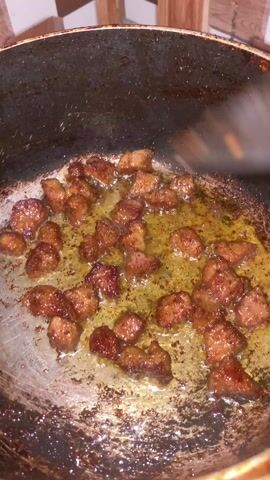 A close-up video shows diced meat frying in oil in a well-used, scratched frying pan. A black cooking utensil, possibly a spatula or spoon, is used to move the meat around in the hot oil, ensuring even cooking. The meat appears to be browning nicely, indicating it is cooking thoroughly. The background features a checkered pattern wall, suggesting a kitchen setting. The handle of the frying pan is visible, indicating it is being held or on a stovetop.