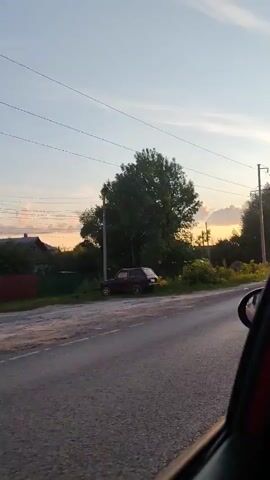 The video captures a view from inside a moving car, looking out at a rural road in Bor, Russia. The scene unfolds during the golden hour, likely late afternoon or early evening, as evidenced by the warm, soft sunlight bathing the landscape. The sky is a clear, pale blue with a few wispy clouds catching the sun's glow.

On the left side of the road, a dark-colored SUV is parked. Beyond the SUV, a large, leafy tree dominates the foreground, casting a shadow. Further back, a few houses with visible roofs and some overgrown vegetation suggest a residential area. Power lines stretch across the sky, supported by utility poles, a common sight in rural settings.

The road itself appears to be paved and in good condition, with a visible shoulder covered in grass and weeds. The right side of the road is lined with more trees, some appearing to be birch trees with their characteristic white bark, interspersed with other deciduous trees. The perspective from the car window offers a dynamic view as the car moves forward, blurring the edges of the scene slightly and emphasizing the sense of travel.

There are no people visible in the frame, but the presence of the car and the houses implies human activity and habitation. The overall mood is peaceful and serene, characteristic of a quiet evening drive through the countryside. The lack of any prominent text or signage contributes to the natural, unfiltered feel of the footage. The environment appears to be a typical Russian rural landscape, characterized by open spaces, modest housing, and abundant greenery.