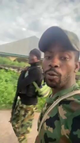 The video shows two individuals in military camouflage outfits walking outside. The person filming is in the foreground wearing a cap and uniform, while the other individual is walking slightly behind and to the side, also in military attire, carrying a rifle and wearing a black top with tactical vest. They appear to be engaged in conversation. The background shows a building, some greenery, and an overcast sky.