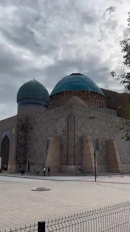 The video shows an exterior view of a building with two domes, one smaller than the other. The building is covered in intricate tilework in shades of blue and white, with geometric patterns. The larger dome is partially covered with scaffolding and what appears to be blue sheeting. Two people are visible walking on a paved courtyard in front of the building. A metal fence runs across the foreground. The sky is overcast with dramatic clouds. Trees are visible on the right side of the frame.