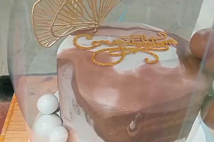 A close-up, slightly blurry shot of a chocolate and white marbled cake. The cake is decorated with a gold "Congratulations" topper and a gold, fan-like ornament. Several white spherical candies or small cakes are visible at the base of the cake on the left side. On the right side, a hand is visible, seemingly adjusting the cake or its protective covering. The background is out of focus, but appears to be an indoor setting with some light colored walls and a wooden surface below the cake.