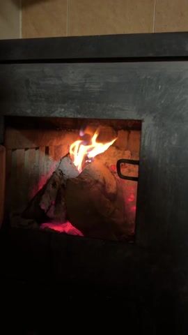 The video shows a close-up view of a fire burning inside a fireplace or wood stove. The flames are primarily orange and yellow, with hints of blue near the base. The fire is burning around and between pieces of wood. The interior walls of the stove appear dark gray. There is some faint audio of people speaking in the background, but the content is unclear.