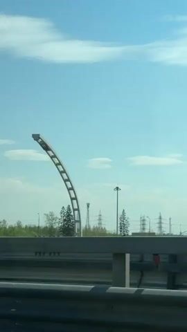 The video shows a view from a vehicle driving on a highway. A grey metal highway divider is visible in the foreground. Beyond the divider, there is a treeline with power lines and a tall, pointed structure in the distance. The sky is blue with scattered white clouds. The camera seems to be mounted in a car, and the movement indicates that the car is moving forward. There is no indication of people visible in the scene.
