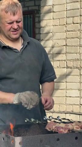 A man in a dark shirt and gloves is grilling meat over an open fire. He is placing pieces of meat onto skewers and arranging them on a grill. The background is a brick wall and some greenery.