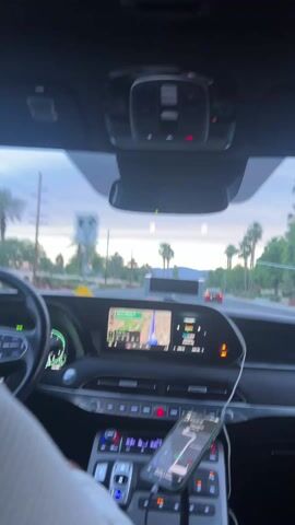 The interior of a car is visible. The car is in motion on a road lined with palm trees and vegetation. The dashboard features an infotainment screen displaying a map. A phone is positioned in the center console, also showing a map. The sky outside is bright, indicating daylight. There are other vehicles visible on the road ahead. The driver’s hand is slightly visible on the steering wheel.