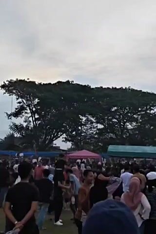 The video shows an outdoor gathering of many people. In the background, there is a large tree and several tents. The tents appear to be set up for an event, with one tent having signage that reads "OPEN TOURNAMENT". The crowd is diverse, with people of various ages and genders, many wearing casual clothing and some wearing headscarves. The ground is covered in grass. The sky is overcast. The overall atmosphere appears to be lively and social, with people milling about and interacting.
