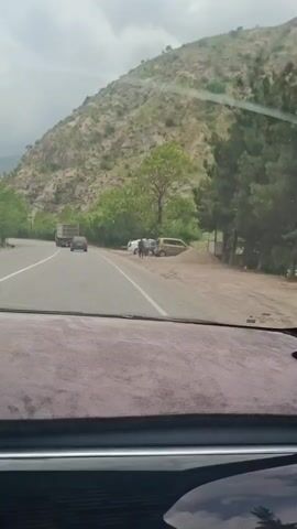 The view from inside a car shows a winding road with a mountain on the right. A large truck and a dark car are driving on the road ahead. On the right side of the road, there are two cars parked near a pile of gravel, and two people are standing near them. The weather appears cloudy. The dashboard of the car with a grey-colored fuzzy cover is visible.