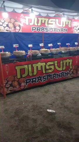 This video shows a dim sum food stall at what appears to be a fair or outdoor market at night. The stall has a red banner with yellow lettering that reads "DIMSUM PRASMANAN". Below this, there is another banner with the same text. Behind the counter, there are several steaming baskets of dim sum lined up on a table. Each basket has a small price tag above it. The ground in front of the stall is covered with dirt or sand. The overall lighting suggests it is evening or night.