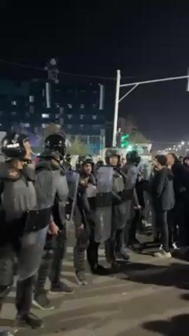 Here is a description of the video:

The video shows a group of riot police standing in a line, facing a group of people. The police are wearing helmets and protective gear, and they are holding shields. The people are standing close to the police line. The scene is taking place at night, with a street and buildings visible in the background.