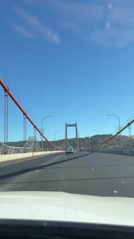 A first-person perspective video shows a clear blue sky with scattered clouds. The camera is positioned inside a vehicle, looking forward through the windshield. The vehicle is driving on a multi-lane bridge. The bridge has orange-colored suspension cables and large white towers. The road surface is asphalt, and there are white lane markings. Several other vehicles, including a white van, are visible on the bridge ahead. The surrounding landscape includes green hills with scattered buildings. The video appears to be captured during the daytime.