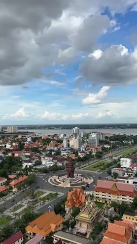 This aerial media file presents a panoramic view of a cityscape, likely Khan Boeng Keng Kang in Cambodia, under a dramatic sky filled with both bright blue patches and voluminous white clouds. The scene is dominated by a large roundabout in the foreground, featuring a prominent brown monument at its center. Surrounding this roundabout is a dense urban landscape characterized by a mix of architectural styles.

Numerous buildings with distinctive orange-tiled roofs, indicative of traditional Cambodian temple architecture, are scattered throughout the city. These are interspersed with modern, multi-story buildings, including some taller high-rise structures visible in the distance. Greenery is also present, with trees lining streets and parks dotting the urban fabric.

A wide river or body of water stretches across the horizon, with more buildings and potentially further urban development on the opposite bank. The presence of vehicles on the roads and the general busyness implied by the dense development suggest that this is an active city. The lighting indicates it is daytime, possibly late morning or early afternoon, given the brightness and cloud cover. The overall impression is one of a vibrant, growing city with a blend of historical and contemporary elements, situated near a significant waterway. No visible text is present in the media file.