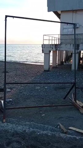 A metal frame stands on a pebbled beach near a building. The frame is composed of vertical and horizontal bars forming a rectangular structure. Two additional bars provide support at the base. In the background is a building supported by concrete pillars over the water. The ocean stretches out to the horizon under a sky showing hints of sunset.