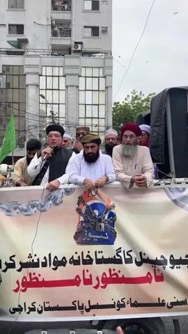 A group of men are standing behind a banner that appears to be protesting. The man on the far left, wearing glasses and a black cap, is speaking into a microphone. He is wearing a black vest over a white shirt. To his right, another man with a beard, wearing a dark cap, is holding prayer beads. A third man, with a beard and wearing a red turban, stands to the right of the second man. Several other men are visible behind them, some wearing caps.

The banner features a logo with a padlock and chains superimposed on it. There is text written in Urdu on the banner. The top line appears to say "جیو چینل کا گستاخانہ مواد نشر کر" which translates to "Geo Channel is broadcasting offensive material". Below this, in red, is "نامنظور نامنظور" meaning "unacceptable, unacceptable". The bottom line reads "سنی علماء پاک پاکستان کراچی" which translates to "Sunni Ulema Pak Pakistan Karachi".

The scene appears to be outdoors, possibly a street, with a building in the background. The sky is overcast, suggesting a cloudy day. The overall mood seems to be one of protest and disapproval. The time of day is not explicitly indicated but the lighting suggests daytime. The location is identified as Karachi Division, Pakistan.