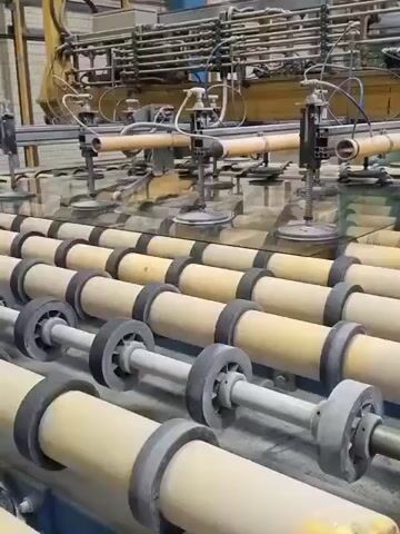 The video shows a glass manufacturing facility. A large pane of glass is being moved along a conveyor belt system. The belt is composed of multiple rows of rollers. Above the glass, a machine with suction cups is attached to the glass, presumably to lift or move it. The overall environment appears to be a factory floor with industrial equipment.