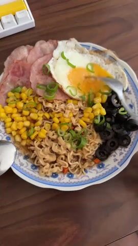 The video shows a plate of food on a wooden table. The plate contains instant noodles, a fried egg, bacon, corn, black olives, and chopped green onions. A fork is lifting some of the noodles from the plate. A spoon is also visible on the left side of the plate. A portion of a white keyboard with yellow and purple keys is visible in the upper left corner of the frame. The lighting appears to be natural light, casting shadows on the table. The overall scene is a close-up, overhead shot of the meal.