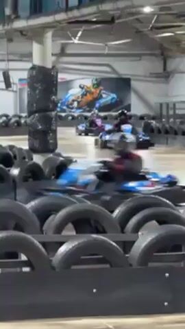 The video shows three go-karts racing around an indoor track. The track is bordered by barriers made of tires and wood. The go-karts are brightly colored with black seats and roll cages. The first kart is blue and white, the second is pink and purple, and the third is mostly black. A large poster depicting a go-kart race is visible in the background. The indoor space is well-lit by overhead lights.