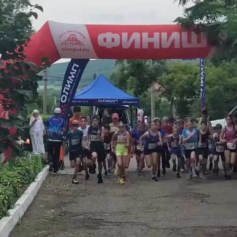 A group of young people is running towards a finish line arch. The arch is red and white with Russian text that says "FINISH" and the logo of "OLIMP." There are tents set up behind the runners. Some of the runners are wearing bib numbers. The ground is a dirt path. The environment appears to be outdoors with trees and foliage visible.