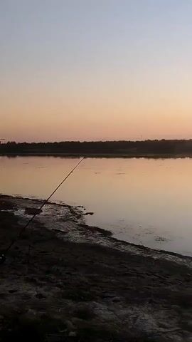 A calm body of water is observed at sunset. The sky transitions from a pale blue at the top to a warm orange and pink near the horizon, reflecting softly on the water's surface. The shoreline in the foreground is dark and somewhat uneven, with sparse vegetation. A fishing rod is visible planted in the ground, extending out over the water on the left side of the frame. Further back, across the water, a silhouetted treeline forms the horizon, punctuated by a few distant structures, including what appears to be a small building with a raised platform. The overall scene is serene and tranquil, capturing the quiet ambiance of twilight.