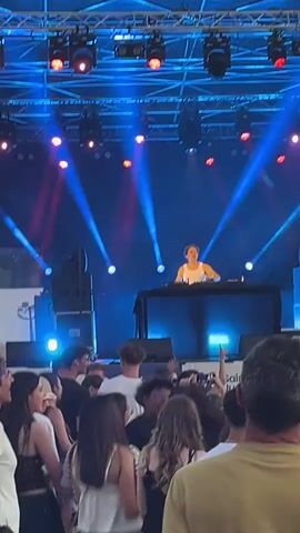 A DJ is performing on a stage at night. The stage is lit with blue and red lights. A crowd of people is gathered in front of the stage, watching the DJ. Some people in the crowd are holding up their phones to record the performance.