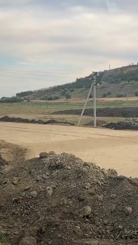 The video captures a construction site on a cloudy day. In the foreground, piles of dirt and construction materials are visible. A dirt road or path is present, with a power pole and overhead wires in the background. Several pieces of construction equipment, including bulldozers and a roller, are operating on the site. One bulldozer appears to be excavating. In the far background, a sloped, treed hill and more distant buildings can be seen. A yellow traffic control box with a red light is visible on the left side of the frame. The overall scene depicts ongoing road or land development work.
