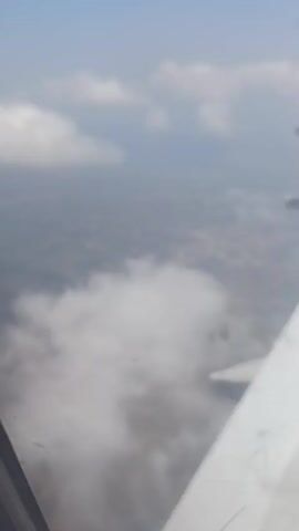 This video is an aerial view from an airplane window, looking down at clouds and land below. An airplane wing is visible on the right side of the frame, with its engines and part of the fuselage. The ground below appears to be a mix of urban areas and greenery, with some bodies of water. The sky is a clear blue with scattered white clouds. The video is a static shot, meaning there is no movement or action depicted other than the implied movement of the airplane.