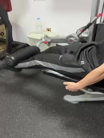 Here is a detailed description of the video. 

The video shows a person using a leg curl machine at a gym. They are wearing black sweatpants with a gray stripe, a black hoodie, and black shoes. The person is seated on the machine and pulling the padded bar toward their body to curl their legs. The machine is silver with a black padded bar. The gym has a black rubber floor, white walls, and a trash can, water bottle, and other gym equipment are visible in the background.