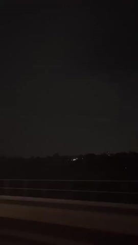 The video shows a dark, possibly night scene from a moving vehicle. In the foreground, there is a horizontal railing and a dark road surface. In the distance, a dark silhouette of trees or a treeline is visible against a very dark sky, with a few faint lights scattered along the horizon, suggesting distant buildings or streetlights. The overall impression is of driving through a rural or suburban area at night.