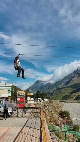 A person is zip-lining across a river with mountains in the background. The person is wearing a white shirt, dark pants, and a harness. The zip line is suspended between two cables. The river is wide and appears to be fast-moving. The mountains are tall and rugged, with some snow at the peaks. The sky is blue with some clouds.