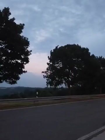 The video shows a moving camera perspective, possibly from a vehicle, traveling along a road. On the left side of the road are large, silhouetted trees, their leaves dark against the twilight sky. The sky itself is a gradient of deep blue and grey with some lighter patches, suggesting it is either early morning or late evening. Beyond the road and a metal guardrail, a landscape of rolling hills is visible, also appearing dark and hazy in the low light. The road surface is asphalt and appears to be a two-lane road with a white line marking the edge. The overall atmosphere is calm and serene, with the muted colors and the natural scenery creating a peaceful ambiance. There are no people or other significant objects or actions depicted in the video.