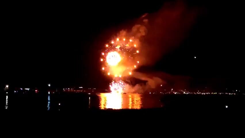 This is a nighttime video of a fireworks display over a body of water. Several large fireworks are exploding in the sky, emitting bright orange and red lights. The explosions are reflected on the dark water, creating shimmering patterns. In the distance, a cityscape is visible with scattered lights. The sky is dark, with the exception of the fireworks and a few faint stars. There are no visible people or boats in the immediate foreground, though distant lights suggest a populated area along the shore. The fireworks are the primary focus of the video, with their colorful bursts dominating the frame.